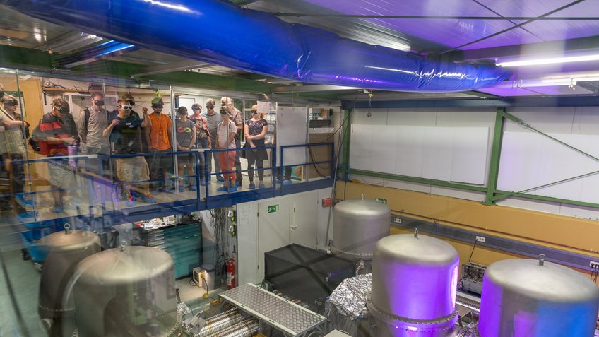 Visit to the GEO600 central building during the Open Day 2018. A group of people are standing on a platform in a laboratory, looking at several large metal tanks and technical devices illuminated by bright lights.
