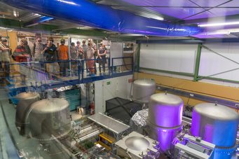 Besichtigung des Zentralgebäudes von GEO600 beim Tag der Offenen Tür im Jahr 2018. Eine Gruppe von Menschen steht auf einer erhöhten Plattform in einem Labor und betrachtet mehrere große Metalltanks und technische Geräte, die von hellen Lichtern beleuchtet werden.