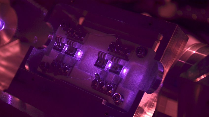 The light from the new laser system recently installed at GEO600 shines through four cubes made of a special crystalline material. Inside these cubes, the properties of a part of the laser light are slightly modified to operate the gravitational-wave detector. The near-infrared (and therefore invisible) laser light appears violet in the image because the infrared filter was removed from the camera used to take the picture. Close-up of an optical device illuminated by purple light.