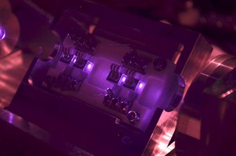 Das Licht des kürzlich bei GEO600 installierten neuen Lasersystems strahlt durch vier Würfel aus einem speziellen Kristallmaterial. In ihnen werden die Eigenschaften von einem Teil des Laserlichts für den Betrieb des Gravitationswellen-Detektors leicht verändert. Das nahinfrarote (und damit eigentlich unsichtbare) Laserlicht erscheint im Bild violett, weil in der zur Aufnahme verwendeten Kamera der Infrarotsperrfilter ausgebaut wurde. Nahaufnahme eines optischen Geräts, beleuchtet in lila Licht.