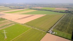 Aerial view of a landscape with large fields in various shades of green and earth colors. Power lines and high-voltage pylons criss-cross the fields. At the lower right edge is a small building complex, which is part of the GEO600 gravitational wave detector. A cityscape is visible in the background.