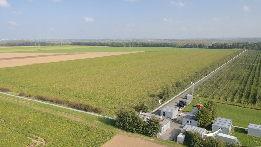 The German-British GEO600 detector south of Hannover, Germany, is a key technology development center of the international gravitational-wave research community. Technologies developed and tested in the GEO project are now used in all large gravitational-wave detectors in the world. Fields stretch to the horizon, with several buildings, two long tunnels at ground level, and a parking lot in the foreground, surrounded by green spaces.