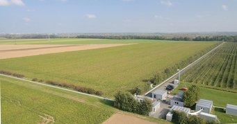 The German-British GEO600 detector south of Hannover, Germany, is a key technology development center of the international gravitational-wave research community. Technologies developed and tested in the GEO project are now used in all large gravitational-wave detectors in the world. Fields stretch to the horizon, with several buildings, two long tunnels at ground level, and a parking lot in the foreground, surrounded by green spaces.
