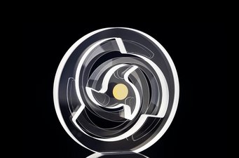 The monolithic glass resonator with paper-thin leaf springs is manufactured using a special joining process. Transparent circular object with intricate internal geometric patterns, including a central yellow dot, set against a dark background.