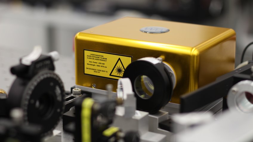 The Ten64 laser source, which was developed at the Max Planck Institute for Gravitational Physics and at Leibniz University Hannover and is commercially offered by VM Photonics GmbH. Photo of a laser source in a golden-yellow housing.