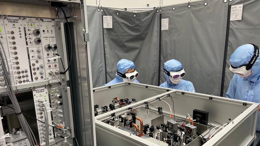 Researcher set up the laser source from Hannover at its new location at ETpathfinder. Three persons wearing blue cleanroom clothing and laser goggles stand behind a complicated experimental setup.