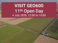 Aerial picture of a landscape with many fields and an L-shaped structure. At its vertex are several buildings and a small parking lot. On top of the image is a red box with text in white letters reading “Visit GEO600, 11th Open Day, 4 July 2026, 12:00 to 16:00”