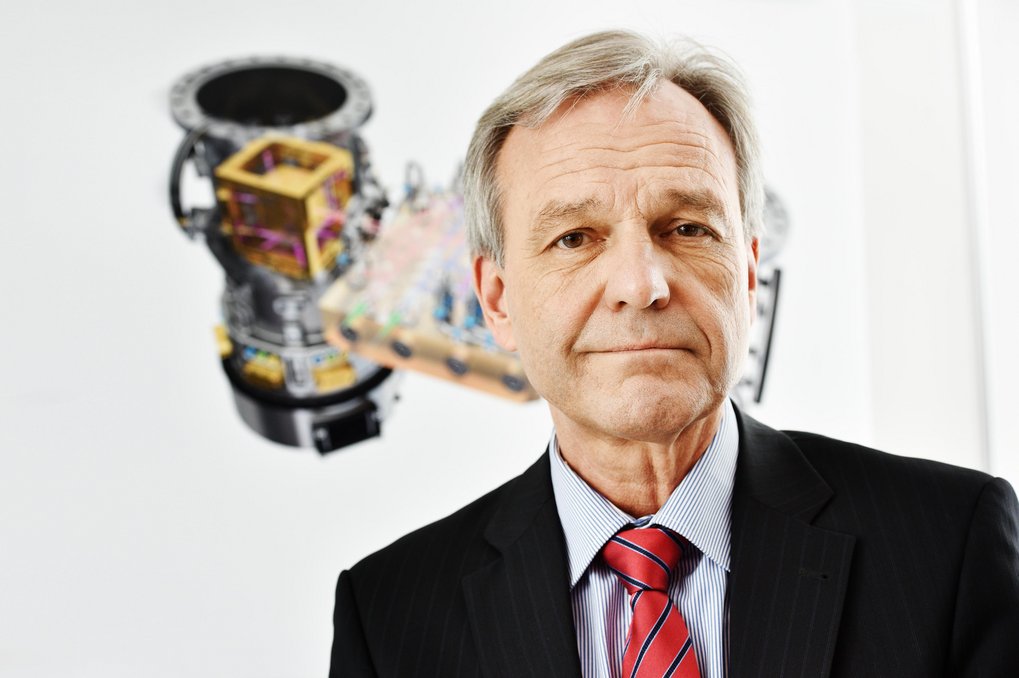 Portrait photo of a man in a black suit, light-blue shirt and a red necktie with blue stripes. In the background is a technical drawing on white.