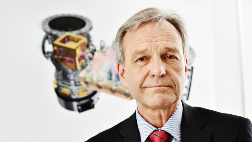Karsten Danzmann, Director Emeritus at the Max Planck Institute for Gravitational Physics (Albert Einstein Institute) Portrait photo of a man in a black suit, light-blue shirt and a red necktie with blue stripes. In the background is a technical drawing on white.