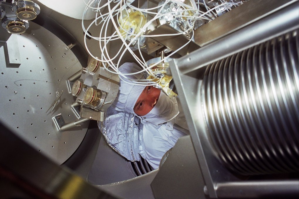 The head of a person in cleanroom clothing in between various metal and glass components.